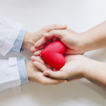 Zwei Menschen halten gemeinsam ein rotes Herzsymbol in ihren Händen – Symbol für Vertrauen, medizinische Fürsorge und das Arzt-Patienten-Verhältnis.